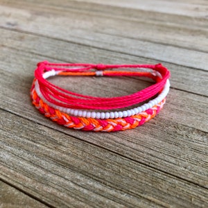 Può includere: Un set di tre braccialetti dell'amicizia colorati con un design intrecciato. I braccialetti sono realizzati con filo e perline rosa, arancioni e bianche.