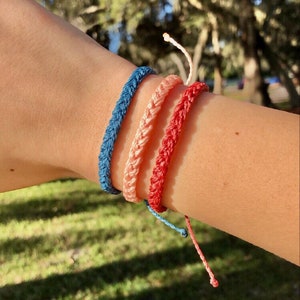 May include: Three braided friendship bracelets in blue, pink, and red colors. The bracelets are worn on a person's wrist.
