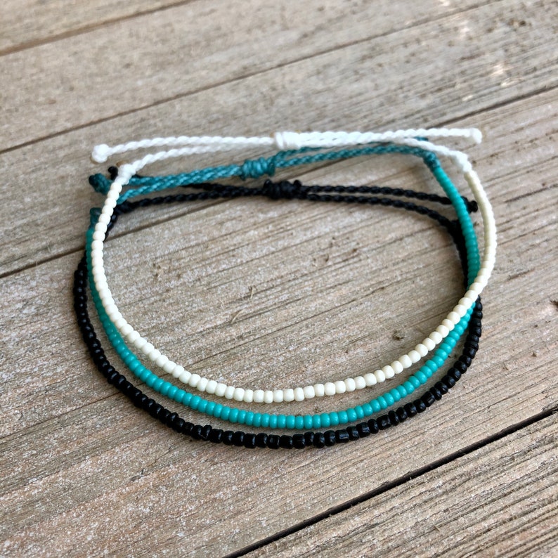May include: Three beaded bracelets in white, turquoise, and black, arranged on a wooden surface. Each bracelet features a string of small, colorful beads. The bracelets are knotted at the ends for adjustable sizing.