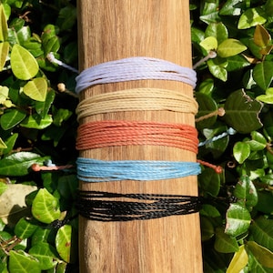 May include: Five colourful bracelets made of woven string, stacked on a wooden surface. The bracelets are in shades of lavender, beige, coral, light blue, and black.