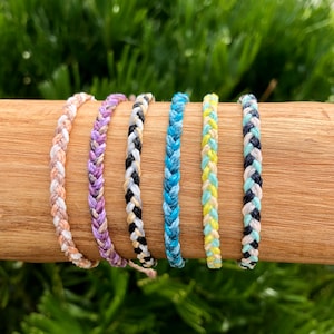 May include: Six colorful braided bracelets made of thin string. The bracelets are in shades of peach, purple, black and white, blue, and yellow and white.