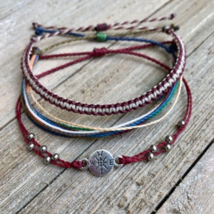 Könnte beinhalten: Satz von fünf bunten Freundschaftsarmbändern mit einem Kompass-Anhänger. Die Armbänder sind aus geflochtener Schnur und in verschiedenen Farben gehalten, darunter Rot, Weiß, Blau, Grün und Braun. Der Kompass-Anhänger ist silberfarben und hat eine schwarze Kompassrose.