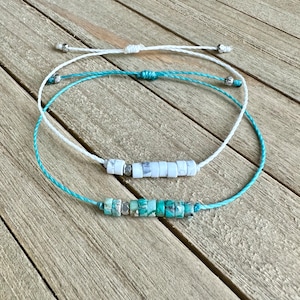 Può includere: Due braccialetti con perline bianche e turchesi su una superficie di legno. I braccialetti sono realizzati con filo bianco e turchese.