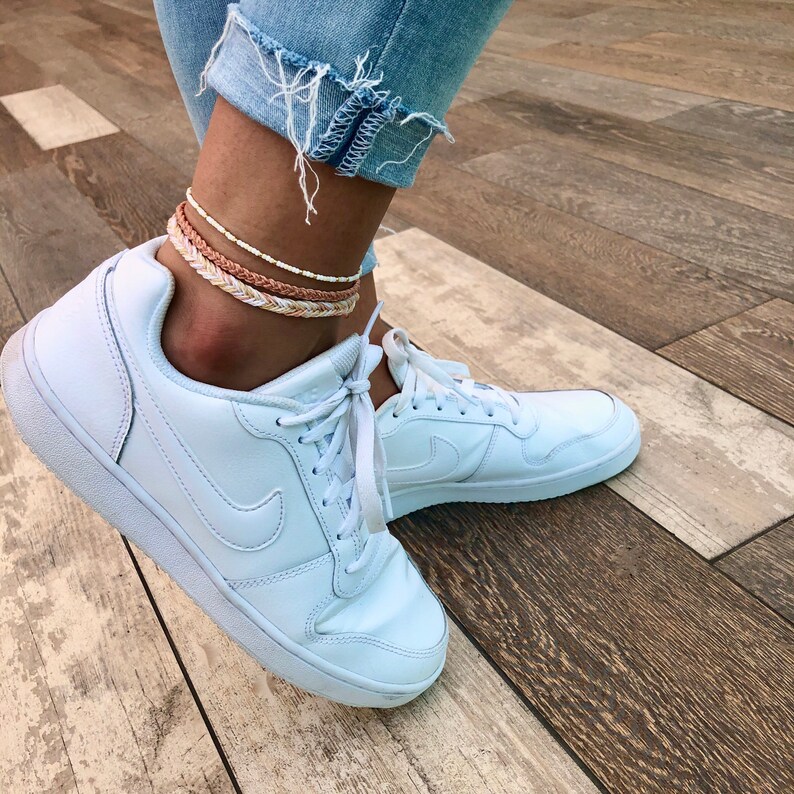 Puede incluir: Un par de zapatillas blancas con cordones blancos y un logotipo Nike blanco. La persona lleva tres tobilleras, una blanca, una marr&oacute;n y otra con un patr&oacute;n blanco y marr&oacute;n.