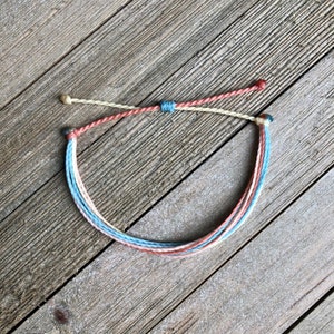 以下が含まれることがあります： 薄い青、桃色、クリーム色のカラーリングで編まれたマルチカラーのブレスレット。ブレスレットは端が結び目になっています。