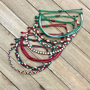 Puede incluir: Una colección de pulseras trenzadas y anudadas de colores rojo, verde y blanco. Algunas pulseras presentan cuentas doradas en forma de estrella y pequeñas cuentas rojas y doradas. Las pulseras están dispuestas sobre una superficie de madera marrón claro.