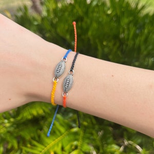 Può includere: Tre braccialetti dell'amicizia con un ciondolo a forma di pallone da calcio argentato. I braccialetti sono arancioni, blu e neri.
