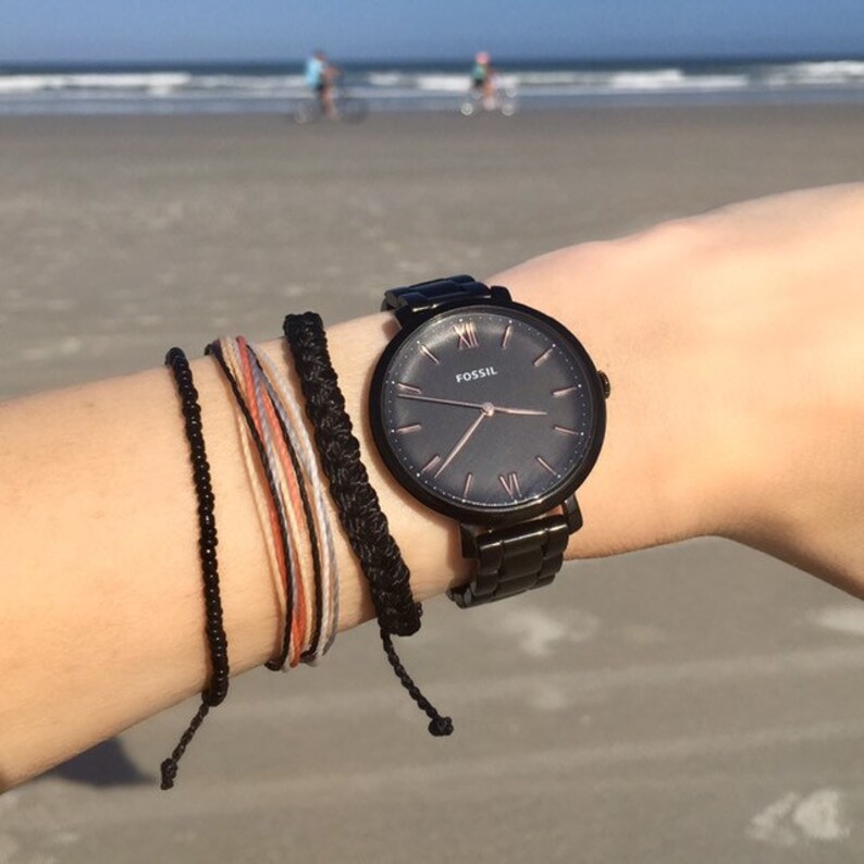 May include: A black Fossil watch with a dark gray face and rose gold hands and markers. Several bracelets in black, orange, and white are worn on the wrist. The background shows a beach with the ocean and people.