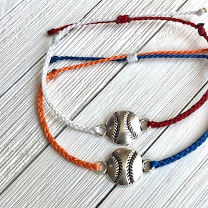 May include: Two friendship bracelets with a silver baseball charm. The bracelets are made with white, red, blue and orange cord.