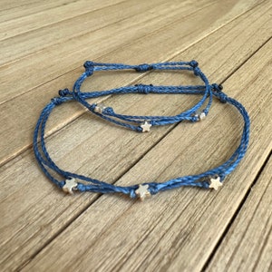 May include: Three blue string bracelets with silver star charms are displayed on a wooden surface. The bracelets are knotted and have a simple, minimalist design. The stars are small and add a touch of whimsy.