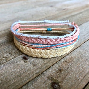 May include: A set of four braided friendship bracelets in shades of pink, white, and beige. The bracelets are layered on top of each other, with the white bracelet featuring small silver beads.