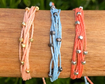 Könnte beinhalten: Drei bunte Freundschaftsarmbänder mit verschiedenen farbigen Schnüren und Metallperlen. Die Armbänder sind zusammengebunden und auf einer Holzoberfläche ausgestellt.