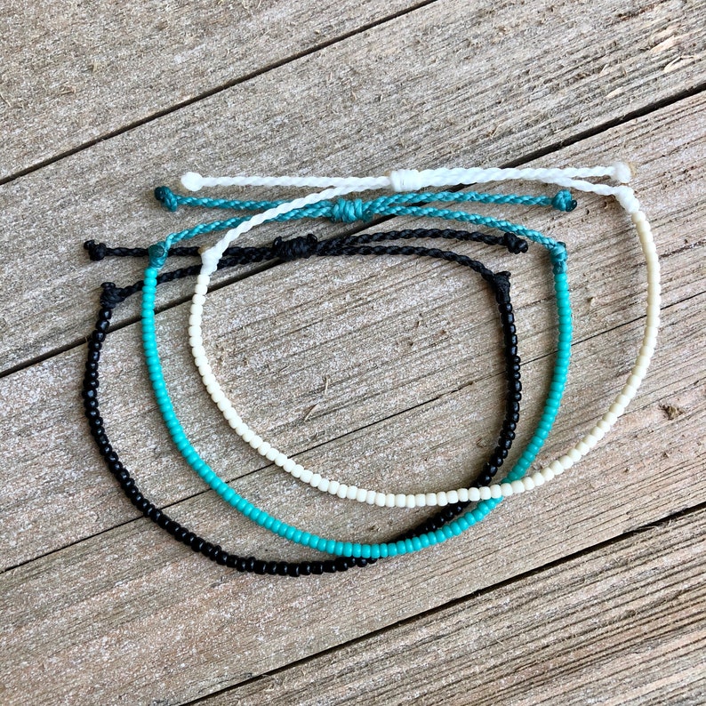May include: Four beaded bracelets in black, turquoise, and white, with adjustable knotted closures. The bracelets are arranged on a weathered wooden surface, showcasing their colors and textures. The bracelets are a simple, minimalist design.