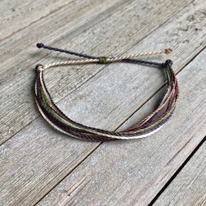 May include: A multi-strand bracelet with a dark brown, cream, green, and burgundy color scheme. The bracelet is made of thin, braided cords and has a simple knot closure.