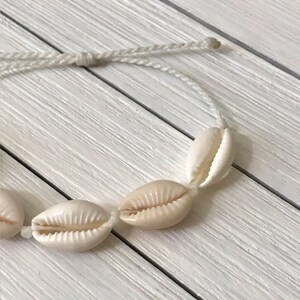 May include: A white string bracelet with five white seashells attached. The bracelet is on a white wooden surface.