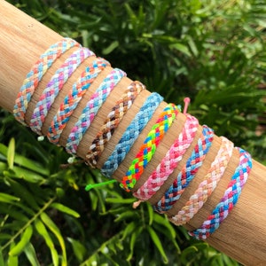 Puede incluir: Una colección de pulseras de amistad trenzadas de colores en varios patrones y colores, incluyendo rosa, azul, blanco, naranja y verde. Las pulseras se muestran sobre una superficie de madera.