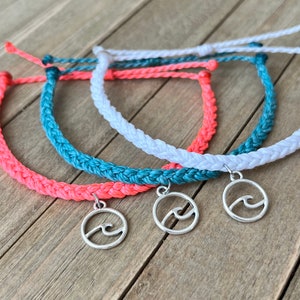 Puede incluir: Tres pulseras de amistad trenzadas con un dije de ola plateado. Las pulseras son de color coral, azul verdoso y blanco.