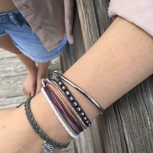 May include: A collection of bracelets in various styles and colors. The bracelets include a silver bangle, beaded bracelets, and woven cord bracelets. One bracelet features a silver leaf charm. The bracelets are stacked on a wrist.