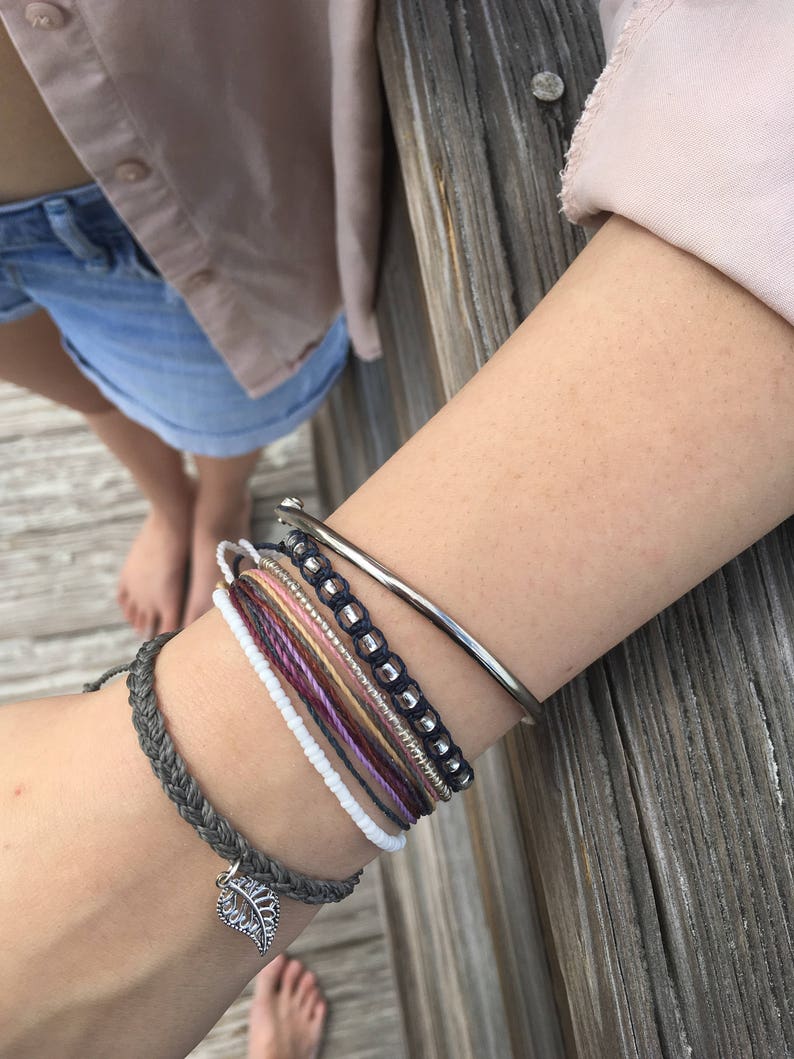 May include: A collection of bracelets in various styles and colors. The bracelets include a silver bangle, beaded bracelets, and woven cord bracelets. One bracelet features a silver leaf charm. The bracelets are stacked on a wrist.