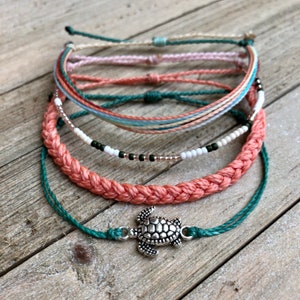 Puede incluir: Un conjunto de cinco pulseras con una variedad de colores y texturas, que incluyen una pulsera de coral trenzada, una pulsera de cuentas con cuentas blancas y negras, y una pulsera azul turquesa con un dije de tortuga plateada.