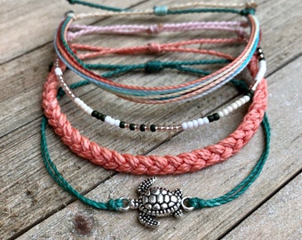 May include: A set of five bracelets featuring a variety of colors and textures, including a braided coral bracelet, a beaded bracelet with white and black beads, and a turquoise blue bracelet with a silver turtle charm.