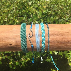 May include: A collection of five bracelets in shades of blue and green. One bracelet is a woven teal color, another is a light blue string, and a third is a braided turquoise. A silver-colored fish hook charm is also included.