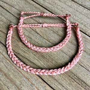 May include: Two pink braided friendship bracelets, one larger than the other, on a wooden surface.
