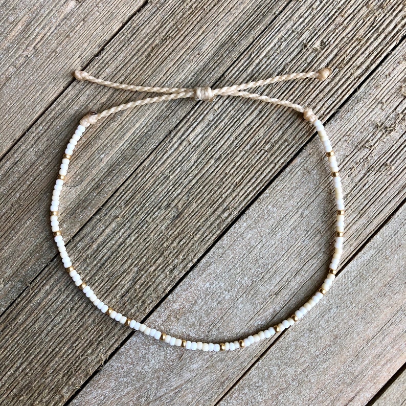 Puede incluir: Un brazalete de cuentas blancas y doradas con cierre de cord&oacute;n anudado. El brazalete est&aacute; hecho con cuentas peque&ntilde;as y redondas y tiene un dise&ntilde;o simple y minimalista.