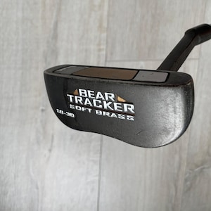 May include: A black mallet-style golf putter featuring a dark finish and a metallic insert on the top face. The head is engraved with the text BEAR TRACKER SOFT BRASS and SB-30 in white lettering, attached to a black shaft extending from the heel.