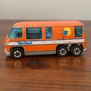 May include: Orange toy van with blue tinted windows, a white stripe, and the words "Palm Beach" on the side. A yellow sun with a palm tree graphic is on the side. The van has six black wheels.