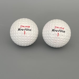 May include: Two white Spalding Kro-Flite golf balls with the number 1 in red. The golf balls have a dimpled surface and the Spalding logo in red and black. The golf balls are on a gray background.