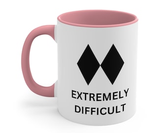 May include: White ceramic mug with a pink interior and handle. The mug features a black diamond symbol and the words "EXTREMELY DIFFICULT" in black, indicating a challenging ski slope.