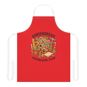 May include: A red apron with white straps featuring a gingerbread house design. The design includes gingerbread people, tools, and the text "GINGERBREAD ENGINEERING TEAM". The apron is designed for kitchen or craft use.