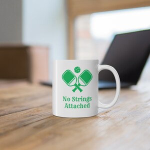 Könnte beinhalten: Weiße Keramik-Kaffeetasse mit einem grünen Pickleball-Schläger- und Ball-Motiv und dem Text "No Strings Attached".