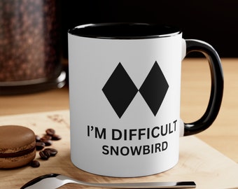 May include: White coffee mug with a black rim and handle. The mug has a black diamond symbol and the text "I'M DIFFICULT SNOWBIRD".
