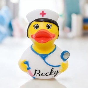 May include: A yellow rubber duck wearing a white nurse's uniform, a white cap with a red cross, and a blue stethoscope. The duck has a red beak and the name "Becky" is written on the front.