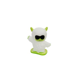 Puede incluir: Una figura blanca de dibujos animados con gafas de sol negras, cuernos verdes y una base verde. La figura tiene los brazos extendidos y una cara sonriente. El juguete est&aacute; dise&ntilde;ado para parecerse a un yeti o monstruo.