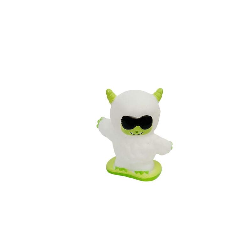 Puede incluir: Una figura blanca de dibujos animados con gafas de sol negras, cuernos verdes y una base verde. La figura tiene los brazos extendidos y una cara sonriente. El juguete est&aacute; dise&ntilde;ado para parecerse a un yeti o monstruo.
