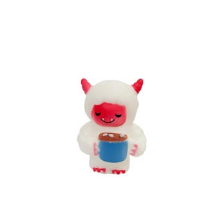 Puede incluir: Peque&ntilde;a figura blanca de un yeti de dibujos animados con cuernos y cara rojos, sosteniendo una taza azul con la parte superior marr&oacute;n. El yeti tiene una expresi&oacute;n sonriente y lleva un abrigo esponjoso.