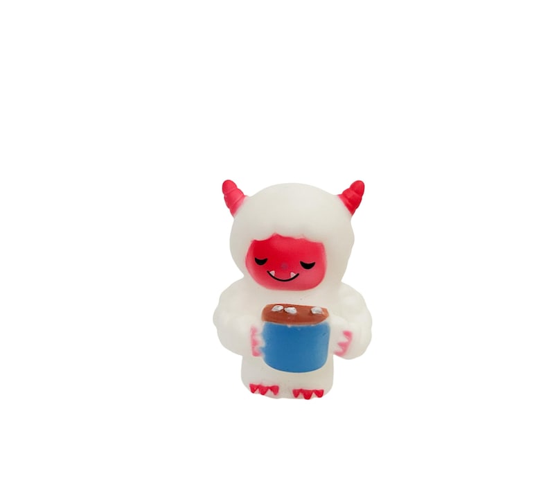 Puede incluir: Peque&ntilde;a figura blanca de un yeti de dibujos animados con cuernos y cara rojos, sosteniendo una taza azul con la parte superior marr&oacute;n. El yeti tiene una expresi&oacute;n sonriente y lleva un abrigo esponjoso.