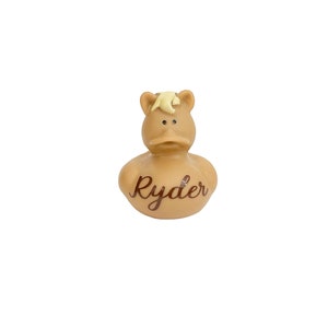 May include: A tan rubber duck with a horse-like appearance, featuring a light yellow mane and the name "Ryder" in a cursive font. The duck has a classic duck bill and small, dark eyes, set against a white background.