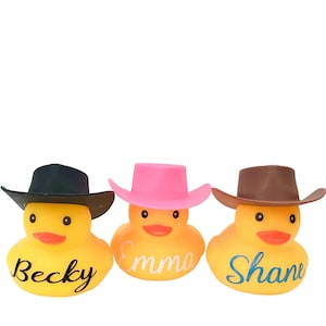 May include: Three yellow rubber duckies wearing cowboy hats. The hats are black, pink, and brown. The ducks are labeled "Becky", "Emma", and "Shane".
