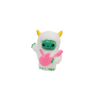 Puede incluir: Una figura de yeti blanca y esponjosa con una cara verde y cuernos amarillos. El yeti sostiene una guitarra el&eacute;ctrica rosa. La figura tiene una cara sonriente y posa como si estuviera tocando la guitarra.