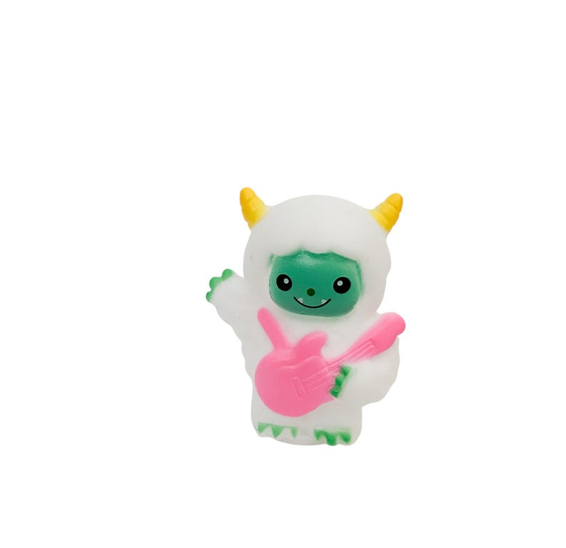 Puede incluir: Una figura de yeti blanca y esponjosa con una cara verde y cuernos amarillos. El yeti sostiene una guitarra el&eacute;ctrica rosa. La figura tiene una cara sonriente y posa como si estuviera tocando la guitarra.