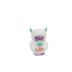 Puede incluir: Una figura de yeti blanco de estilo caricatura, con cuernos verdes y una cara morada, sosteniendo un helado colorido. El helado tiene una bola verde, un centro rosa y un cono naranja. El yeti tiene una expresi&oacute;n feliz.
