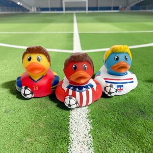 May include: Three rubber duck toys dressed as soccer players. The ducks are yellow, red, and blue, each with a different uniform and hairstyle. Each duck holds a soccer ball. The background is a green field with white lines.