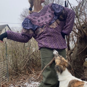 May include: A woman wearing a purple and blue patterned sweater with a ruffled neckline and green trousers is standing in a garden with a dog. The dog is white with brown and black patches and is holding a stick in its mouth.