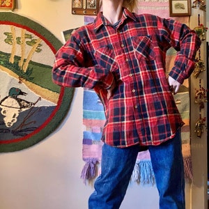 May include: A red and black plaid flannel shirt with a button-down front and two chest pockets. The shirt is worn over a pair of blue jeans.