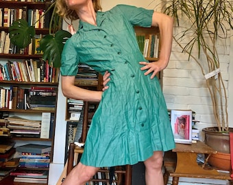 May include: A woman wearing a teal green button-down dress with short sleeves. She is standing in a room with bookshelves and plants.