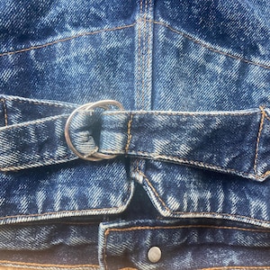 Puede incluir: Primer plano de una chaqueta vaquera azul con cierre de hebilla de metal plateado. La chaqueta presenta un detalle de cinturón con hebilla redonda y extremo puntiagudo. El tejido vaquero tiene un aspecto descolorido y desgastado con costuras visibles.